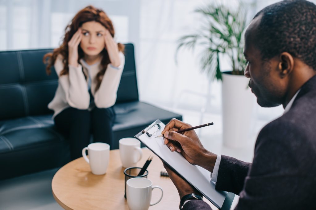Upset woman sitting on sofa and psychiatrist writing in clipboard
